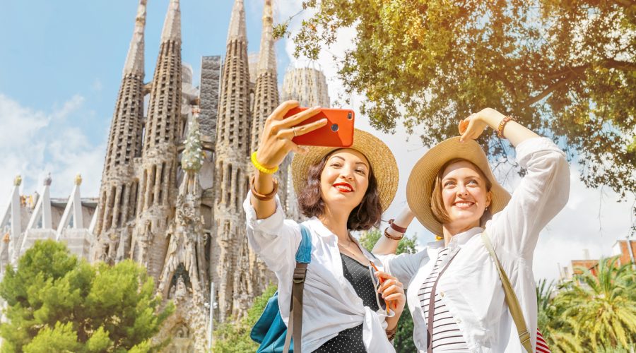 BARCELONA, SPAIN - 11 JULY 2018: Young girls friends making selfie photo on her smartphone in front of the famous Sagrada Familia catholic cathedral. Travel in Barcelona concept